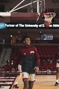 A basketball player in a maroon and white uniform smiles while walking on a basketball court. The ball is mid-air near the hoop, indicating a successful shot. In the background, there are empty red stands and a digital banner displaying text related to the University of Denver Athletics.