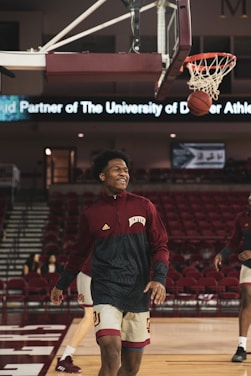 A basketball player in a maroon and white uniform smiles while walking on a basketball court. The ball is mid-air near the hoop, indicating a successful shot. In the background, there are empty red stands and a digital banner displaying text related to the University of Denver Athletics.