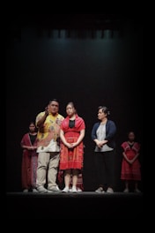 Several individuals are standing on a stage against a dark background, dressed in a mix of traditional and modern clothing. The group is positioned in a way that suggests they are part of a performance or cultural presentation.