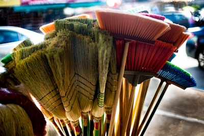 A collection of colorful brooms with varied bristle shapes and materials. They are closely arranged, with some featuring natural, straw-like bristles, while others have synthetic, vibrant-colored bristles like red, blue, and green. The brooms are held together in a bunch, creating a visually striking contrast against a blurred urban background.