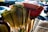 A collection of colorful brooms with varied bristle shapes and materials. They are closely arranged, with some featuring natural, straw-like bristles, while others have synthetic, vibrant-colored bristles like red, blue, and green. The brooms are held together in a bunch, creating a visually striking contrast against a blurred urban background.