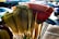 A collection of colorful brooms with varied bristle shapes and materials. They are closely arranged, with some featuring natural, straw-like bristles, while others have synthetic, vibrant-colored bristles like red, blue, and green. The brooms are held together in a bunch, creating a visually striking contrast against a blurred urban background.