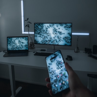 A modern, minimalist workspace features a large monitor, a laptop, and a smartphone, all displaying the same abstract wallpaper with the text 'Hustle Anywhere'. The desk is uncluttered, with a few decorative items and peripherals like a keyboard and mouse. Soft lighting adds to the calm ambiance of the room.