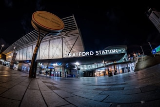 A modern transport station entrance at night, featuring illuminated signage that reads 'Stratford Station.' The area is well-lit with overhead lights and displays a futuristic architectural design with glass and metal elements. The wide-angle view captures the expansive pavement leading up to the entrance, and a round sign indicates 'Welcome to Newham, London.'