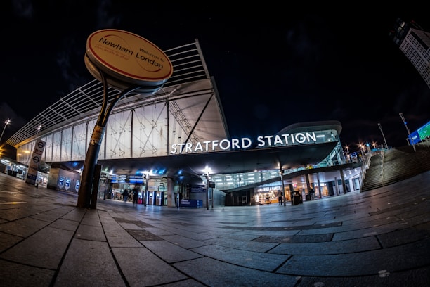 A modern transport station entrance at night, featuring illuminated signage that reads 'Stratford Station.' The area is well-lit with overhead lights and displays a futuristic architectural design with glass and metal elements. The wide-angle view captures the expansive pavement leading up to the entrance, and a round sign indicates 'Welcome to Newham, London.'