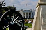 A large white building with a flagpole on top, featuring a U.S. flag waving. The building is set against a background of cloudy skies and green trees. Foreground elements include a large cannon and a stone monument. People are gathered in front of the building, some near a fountain and surrounding greenery.