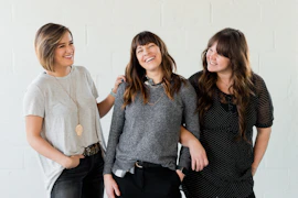 3 women smiling and standing beside white wall