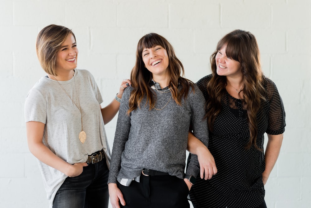 3 mulheres sorrindo e de pé ao lado da parede branca