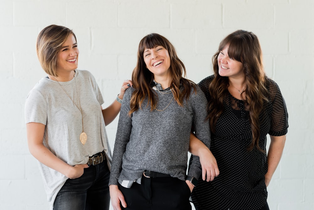 3 femmes souriantes et debout à côté d’un mur blanc