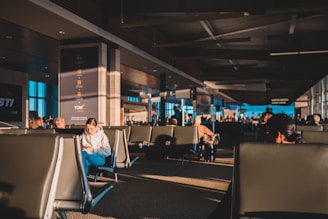 A spacious airport terminal is filled with people sitting in rows of black seats, some engaged with their phones or reading materials. The sun casts warm, elongated shadows across the floor and seats, creating a contrast with the cool tones of the terminal's interior design. Large windows on the side allow natural light to illuminate the area, and a few signs and screens display flight information.