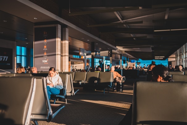 A spacious airport terminal is filled with people sitting in rows of black seats, some engaged with their phones or reading materials. The sun casts warm, elongated shadows across the floor and seats, creating a contrast with the cool tones of the terminal's interior design. Large windows on the side allow natural light to illuminate the area, and a few signs and screens display flight information.