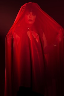 A person with bold makeup and dark lipstick is partially obscured by a sheer red fabric. The lighting is dim, casting a dramatic shadow and highlighting the intensity of the red color.