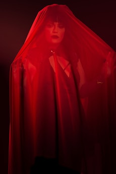 A person with bold makeup and dark lipstick is partially obscured by a sheer red fabric. The lighting is dim, casting a dramatic shadow and highlighting the intensity of the red color.