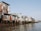 Several dilapidated houses constructed from corrugated metal sheets stand on wooden stilts along a riverbank. The structures appear weathered, with rust and patches of varying colors visible on the walls. The river in the foreground is calm, reflecting the sky and buildings. There is a clear blue sky in the background, suggesting a sunny day.