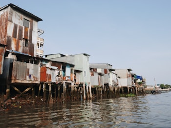Several dilapidated houses constructed from corrugated metal sheets stand on wooden stilts along a riverbank. The structures appear weathered, with rust and patches of varying colors visible on the walls. The river in the foreground is calm, reflecting the sky and buildings. There is a clear blue sky in the background, suggesting a sunny day.