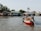 A small boat navigates through calm waters, loaded with various fruits and vegetables. Two people are seated in the boat, suggesting a market or trading activity. In the background, there are several houses on stilts lined along the riverbank, surrounded by greenery.