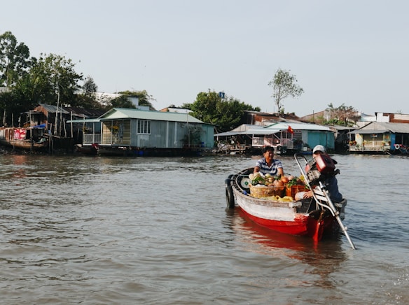A small boat navigates through calm waters, loaded with various fruits and vegetables. Two people are seated in the boat, suggesting a market or trading activity. In the background, there are several houses on stilts lined along the riverbank, surrounded by greenery.
