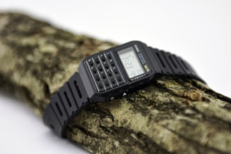 A digital wristwatch with a black strap and calculator buttons is placed on a textured piece of wood. The watch display shows the time and some digital indicators, and the overall setting is minimalistic with a focus on the watch's features.