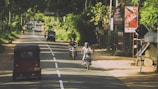 A rural road with vehicles including motorcycles and an auto rickshaw traveling along it. Trees and greenery line the sides of the road, creating a canopy effect. Various signs, including advertisements for Coca-Cola, are visible along the roadside. The scene is bright and sunny.