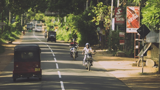A rural road with vehicles including motorcycles and an auto rickshaw traveling along it. Trees and greenery line the sides of the road, creating a canopy effect. Various signs, including advertisements for Coca-Cola, are visible along the roadside. The scene is bright and sunny.