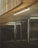 An industrial, dimly lit environment featuring a metal gate under a ceiling with exposed fluorescent lights. The walls have a concrete texture and there are shadows creating a sense of depth. The area beyond the gate is dark and appears enclosed.