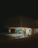A modern building with a large, illuminated storefront and glass windows displaying an array of items inside. The exterior is dark, with a checkered pattern on the ground reflecting some of the light from the interior.
