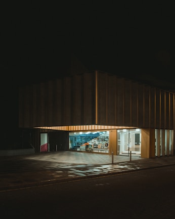 A modern building with a large, illuminated storefront and glass windows displaying an array of items inside. The exterior is dark, with a checkered pattern on the ground reflecting some of the light from the interior.