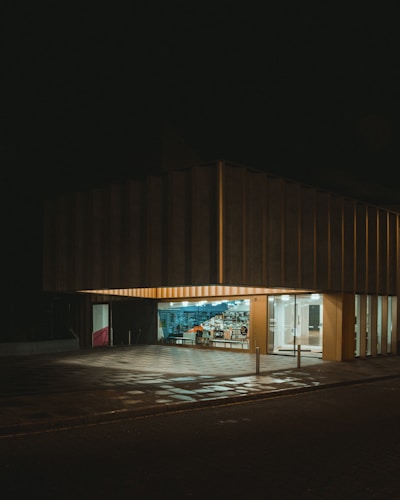 A modern building with a large, illuminated storefront and glass windows displaying an array of items inside. The exterior is dark, with a checkered pattern on the ground reflecting some of the light from the interior.