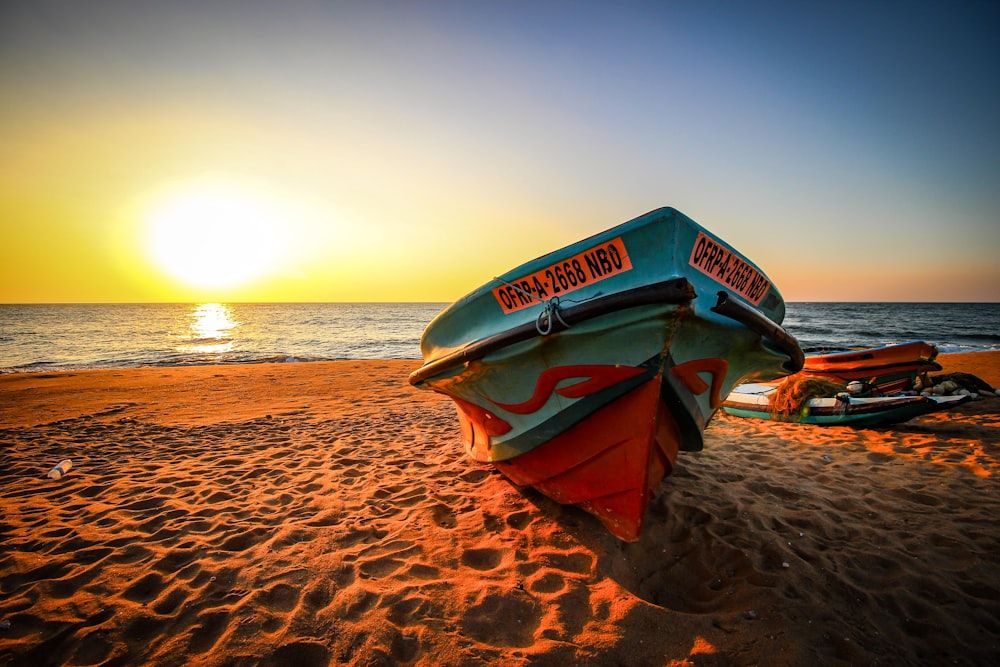 Barco rojo y azul en arena marrón cerca del cuerpo de agua durante la puesta del sol