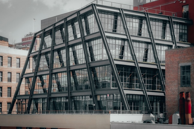 A modern building with a unique angular steel frame structure and glass facade, set among older brick buildings. The architectural design features multiple stories with large windows, prominent beams, and a rooftop view of other urban structures.