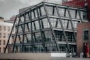 A modern building with a unique angular steel frame structure and glass facade, set among older brick buildings. The architectural design features multiple stories with large windows, prominent beams, and a rooftop view of other urban structures.