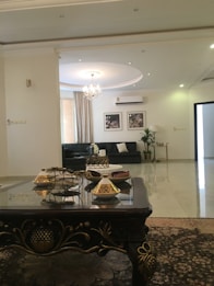 A spacious and elegantly decorated living room with a dark ornate coffee table featuring decorative items. Behind it, there is a dark green sofa with cushions, complemented by a stylish chandelier hanging from the ceiling. The walls are adorned with framed floral artwork and the floor is covered with a patterned rug.