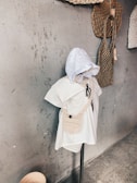 A mannequin dressed in a white outfit with a crocheted bag and a hood stands against a textured gray wall. There are two woven bags hanging on the wall beside it, adding an artisanal feel to the setup.