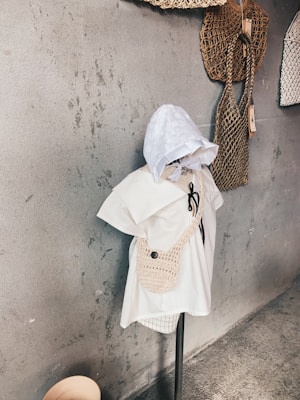 A mannequin dressed in a white outfit with a crocheted bag and a hood stands against a textured gray wall. There are two woven bags hanging on the wall beside it, adding an artisanal feel to the setup.