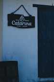 A rustic sign with the words 'Brew House Catarina Ceraria' is hanging on a textured white wall. Next to the sign is a worn, dark blue wooden door, and part of a wooden barrel is visible at the bottom left corner.