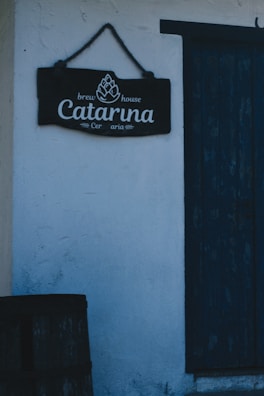 A rustic sign with the words 'Brew House Catarina Ceraria' is hanging on a textured white wall. Next to the sign is a worn, dark blue wooden door, and part of a wooden barrel is visible at the bottom left corner.