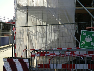 A construction site is partially covered with a translucent tarp, revealing scaffolding behind it. A metal fence with red and white caution tape runs across the foreground. A green safety sign with arrows pointing inward features white silhouettes of people and Dutch text 'VERZAMELPLAATS,' indicating an assembly point.