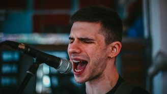 A person passionately singing into a microphone, with eyes closed and an expressive facial expression. The background is slightly blurred, suggesting an indoor setting.