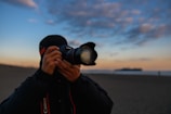 A person is holding a Canon camera, preparing to take a photo on a beach during sunset. The sky is filled with soft, scattered clouds and the horizon shows a distant silhouette of land or islands.