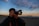 A person is holding a Canon camera, preparing to take a photo on a beach during sunset. The sky is filled with soft, scattered clouds and the horizon shows a distant silhouette of land or islands.