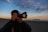 A person is holding a Canon camera, preparing to take a photo on a beach during sunset. The sky is filled with soft, scattered clouds and the horizon shows a distant silhouette of land or islands.