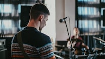 A musician is playing a guitar in a dimly lit room with a microphone stand in the foreground. In the background, another musician is playing drums. The scene includes music equipment and speakers, with light filtering through large windows, creating a relaxed atmosphere.
