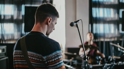 A musician is playing a guitar in a dimly lit room with a microphone stand in the foreground. In the background, another musician is playing drums. The scene includes music equipment and speakers, with light filtering through large windows, creating a relaxed atmosphere.