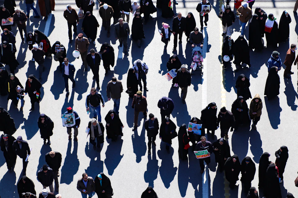 The image depicts a large group of people walking on a street, with their shadows cast onto the pavement below them. The perspective is from above, providing an overhead view of the scene. The crowd consists of individuals dressed in various attire, including coats and hats, suggesting that it might be a cold or overcast day. Some are carrying bags, indicating they may be shopping or running errands. The overall atmosphere conveys a sense of activity and movement as people go about their daily routines.