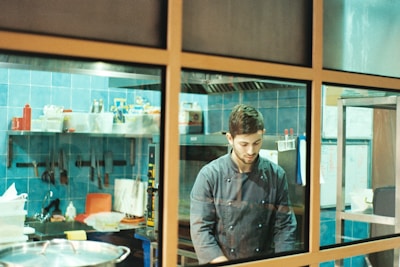 A chef in a dark chef's jacket is working in a kitchen with blue tiled walls. The kitchen is equipped with several shelves that hold various ingredients and cooking tools. Utensils hang on the wall, and a large pot is visible in the foreground. The chef appears focused on his task.
