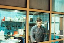 A chef in a dark chef's jacket is working in a kitchen with blue tiled walls. The kitchen is equipped with several shelves that hold various ingredients and cooking tools. Utensils hang on the wall, and a large pot is visible in the foreground. The chef appears focused on his task.