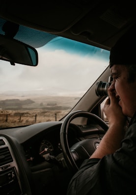 A person is sitting inside a car, using a camera to take a photograph through the window. The interior of the car, including the steering wheel and dashboard, is visible. Outside, there is a landscape featuring a cloudy sky and distant hills or fields.