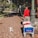 A person wearing a red shirt and gray pants is walking along a brick-paved sidewalk. They are pulling a blue cart labeled 'Beachcomber' with red and white items inside. A small white and tan dog is walking ahead of them on the sidewalk. There are trees and greenery to the left, and the side of a bus and street to the right. Another person wearing a blue outfit is seen walking in the background.