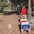 A person wearing a red shirt and gray pants is walking along a brick-paved sidewalk. They are pulling a blue cart labeled 'Beachcomber' with red and white items inside. A small white and tan dog is walking ahead of them on the sidewalk. There are trees and greenery to the left, and the side of a bus and street to the right. Another person wearing a blue outfit is seen walking in the background.
