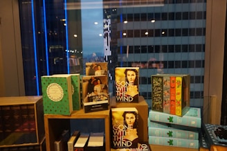 A collection of books is arranged on a display shelf in front of a window with a cityscape view. Dominant books include 'Gone With the Wind' and various colorful, decorative covers. The city buildings outside are lit up with blue and white lights, contributing to the ambient nighttime atmosphere.